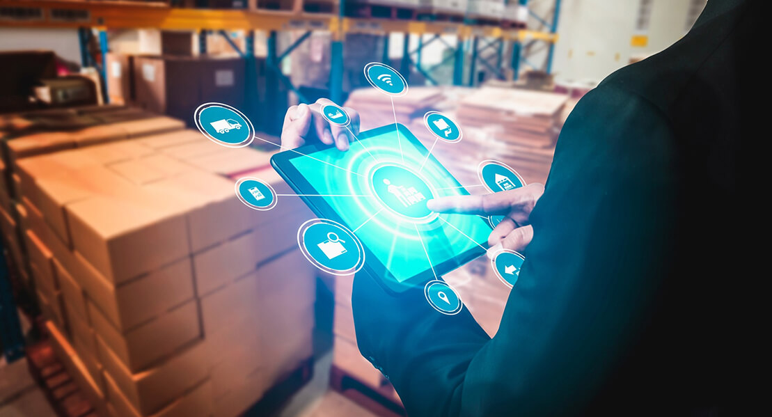 A person in a suit interacts with a tablet displaying logistics icons, set against a backdrop of stacked cardboard boxes in a warehouse.