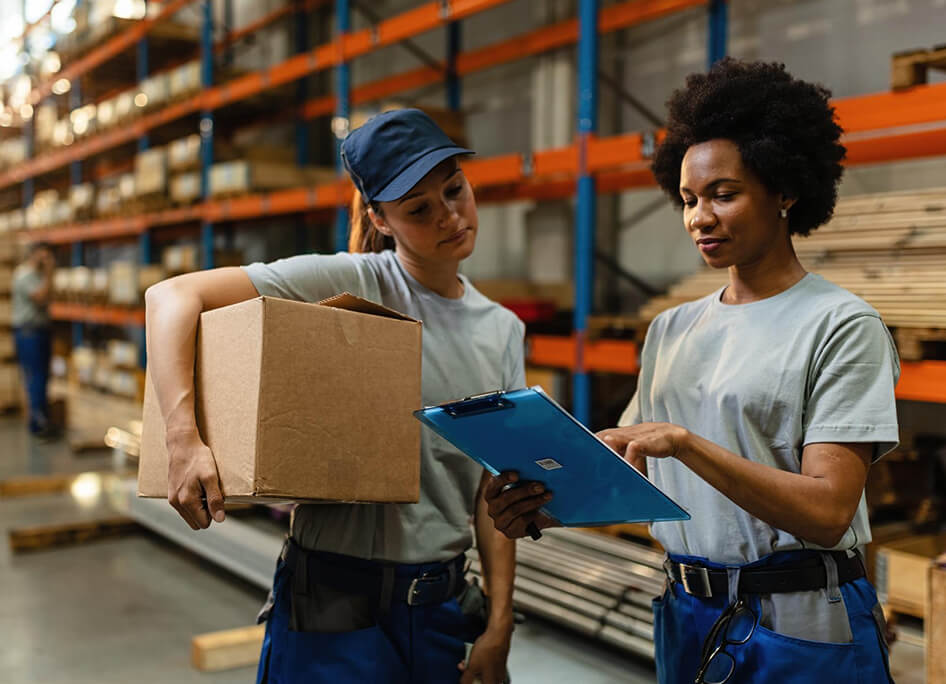 Two warehouse workers discuss inventory, one holding a cardboard box and the other reviewing a clipboard in a busy storage area.