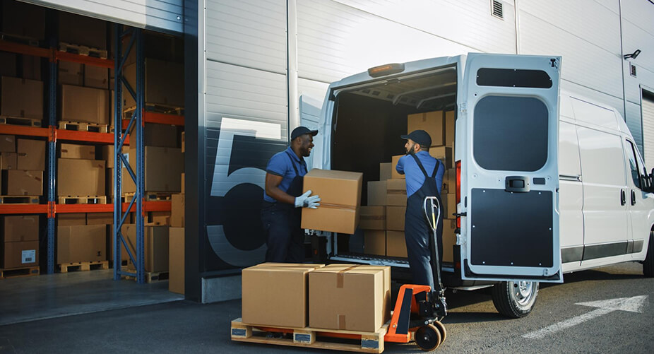 Two workers loading cardboard boxes into a white delivery van outside a warehouse, with stacks of boxes in the background.